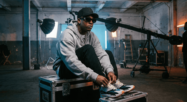 Rapper in a grey hoodie and cap lacing his sneakers on a flight case on a video set