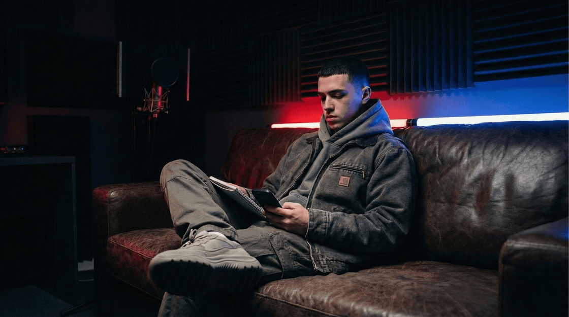 Rapper in a washed workwear jacket writing in a notebook on a leather couch in a studio lounge lit red and blue