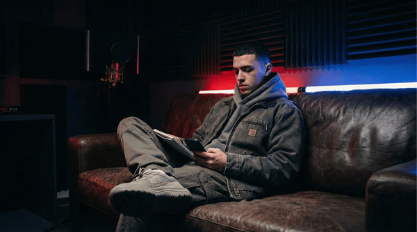 Rapper in a washed workwear jacket writing in a notebook on a leather couch in a studio lounge lit red and blue