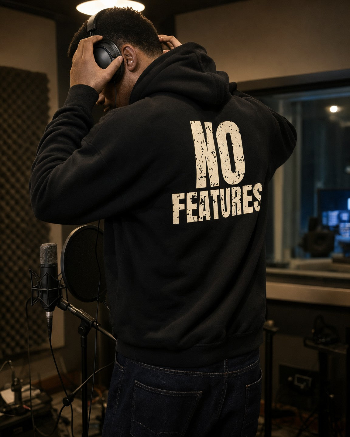 Man wearing the No Features hip hop hoodie in black, back print, pulling on studio headphones in a vocal booth
